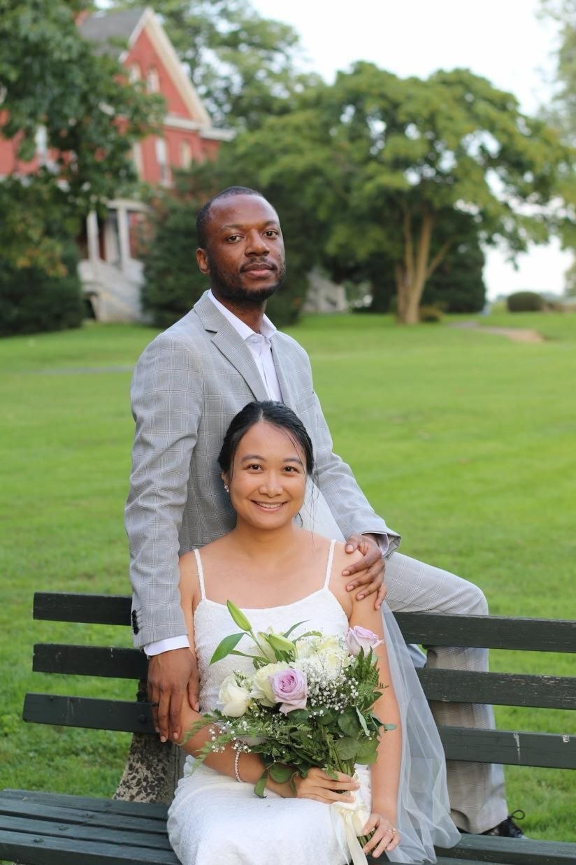 Omar stands behind Krishel, who sits on a park bench with a bouquet.