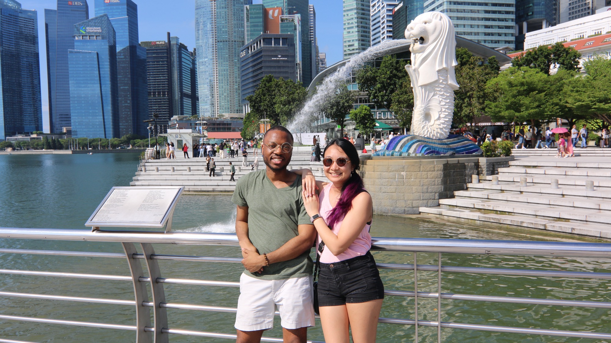 Krishel and Omar stand arm in arm by the water in front of the Merlion in Singapore.